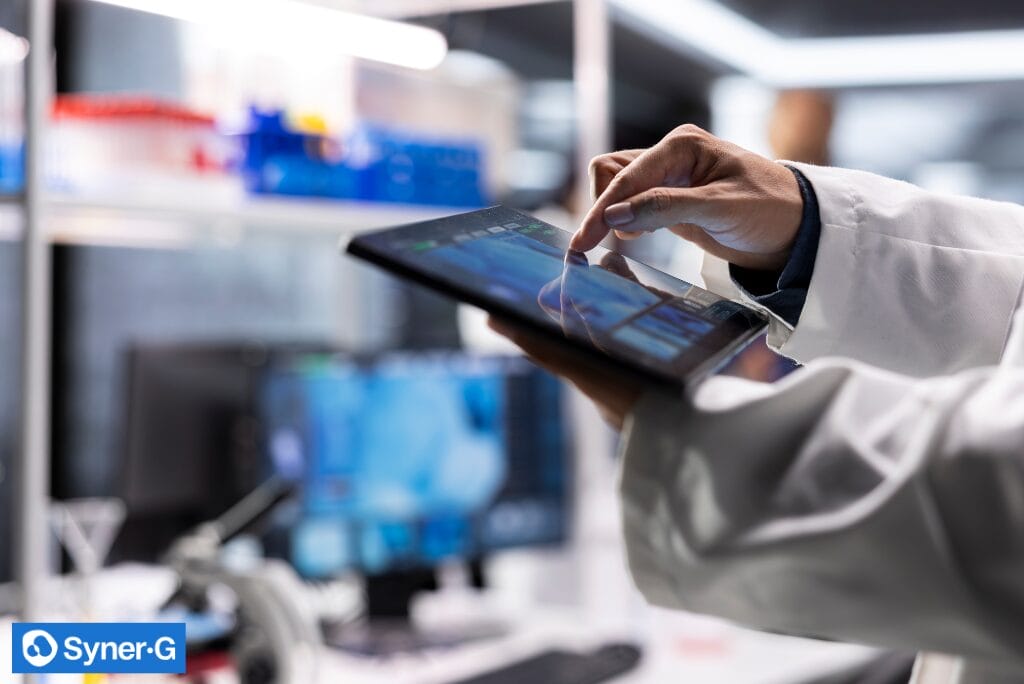 Researcher in a lab coat uses a tablet touchscreen in a laboratory setting to monitor digital data and workflows for clinical trial supply chain operations.