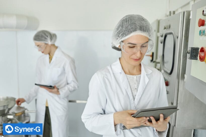 Pharmaceutical manufacturing staff in lab coats, hairnets, and safety glasses use tablets in a cleanroom production area to monitor processes and compliance.
